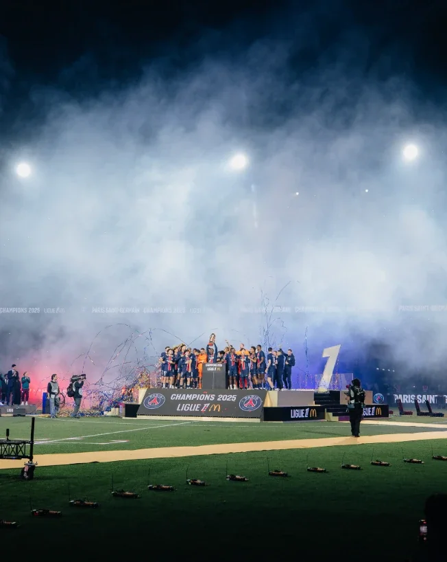 Paris Saint-Germain players lifting the Ligue 1 trophy on a podium, under floodlights and smoke
