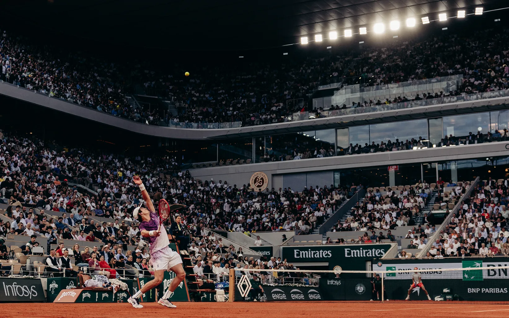 A player stretching for a forehand on the clay of Court Philippe-Chatrier, stands full under the lights